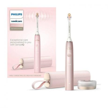 Spazzolino elettrico Philips Sonicare 9900 Prestige: uno spazzolino elettrico rosa con custodia da trasporto abbinata e base di ricarica dorata accanto a una scatola di imballaggio rosa su sfondo bianco