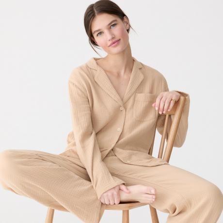 Model kompleta pižame z dolgimi rokavi iz mehke gaze J.Crew, oblečen v komplet pižame iz rjave gaze, sedi na lesenem stolu na belem ozadju