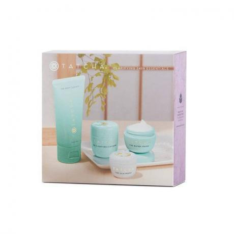 Tatcha Skin Essentials Set: Een doos op een witte achtergrond met een afbeelding van een lichtblauwe buis, twee kleine bijpassende potjes en een klein wit potje zittend op een houten tafel met een voor een raam
