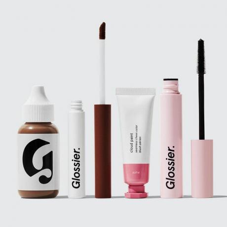 Una foto de los productos Glossier, incluido el tinte Perfecting Skin Tint, las sombras de ojos líquidas Skywash, el rubor en crema Cloud Paint y el rímel Lash Slick sobre un fondo gris